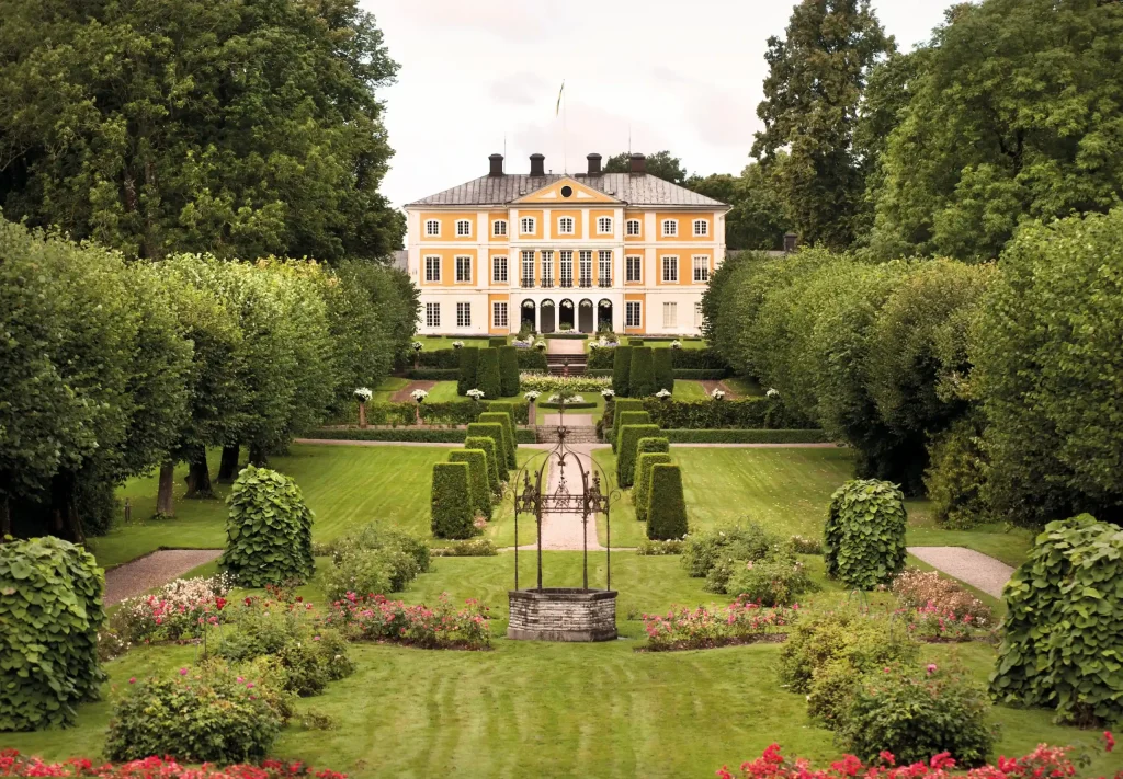 Stor gul herrgård med välordnad symmetrisk trädgård i förgrunden.