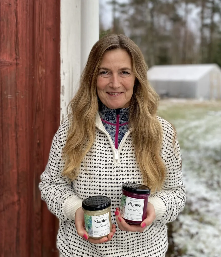 En kvinna i stickad tröja med långt hår stor med två burkar i händerna mot en röd trädvägg, landskap i bakgrunden. 