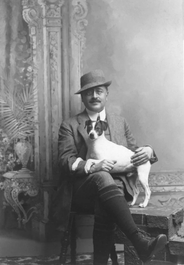 En man i hatt, monokel, mustasch och jaktkläder sitter med en Jack Russell-terrier i sitt knä.