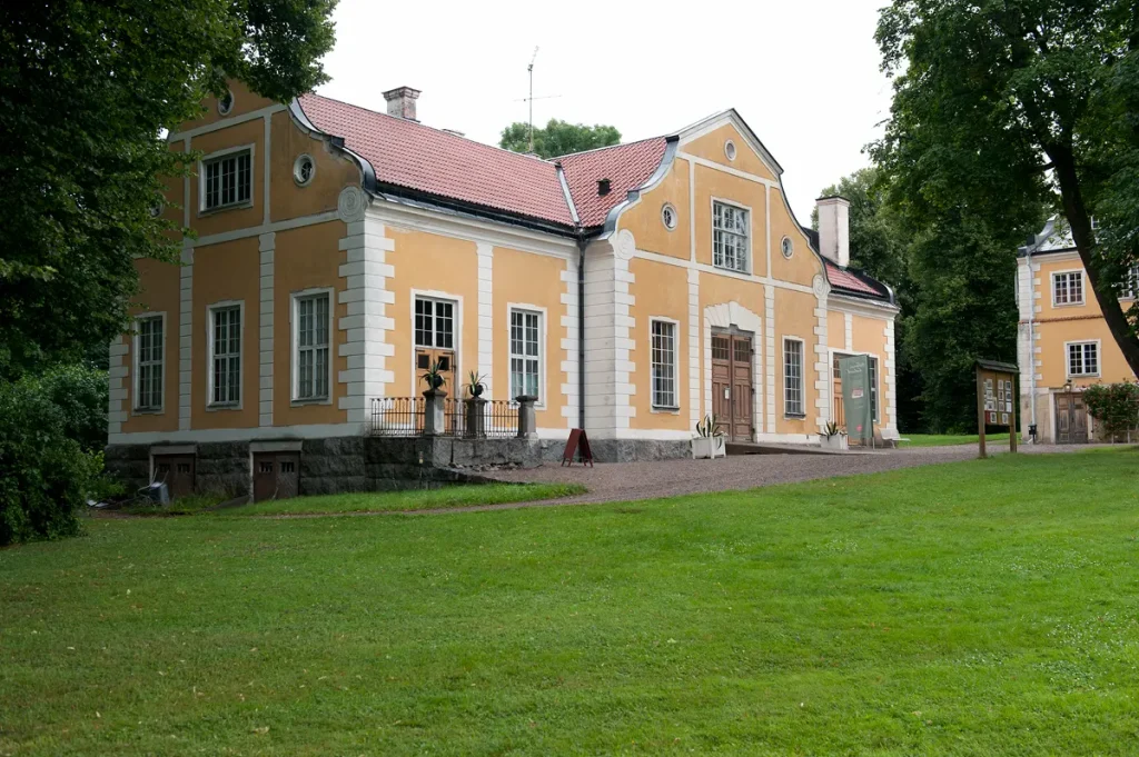 Gul byggnad med vita knutar och svart tak, omgiven av grönskande träd och en trädgård.