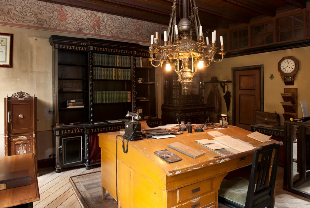 Ett äldre kontor med ett stort slitet skrivbord i mitten, en kristallkrona i taket och en bokhylla på väggen. Golvet är i trä och en stor mjuk matta finns under skrivbordet.