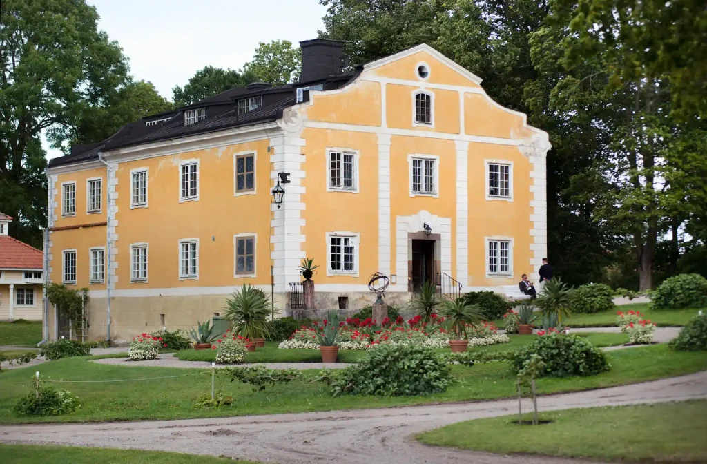 Gul byggnad med vita knutar och svart tak, omgiven av grönskande träd och en trädgård med blommor i förgrunden.