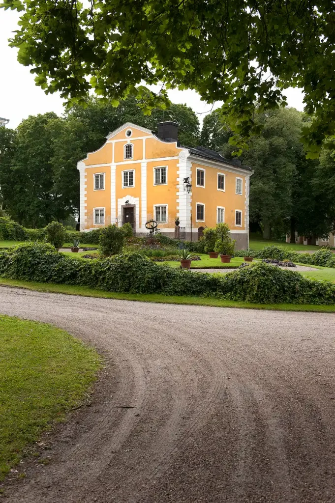Gul byggnad med vita knutar och svart tak, omgiven av grönskande träd samt välordnad trädgård och grusväg i förgrunden.