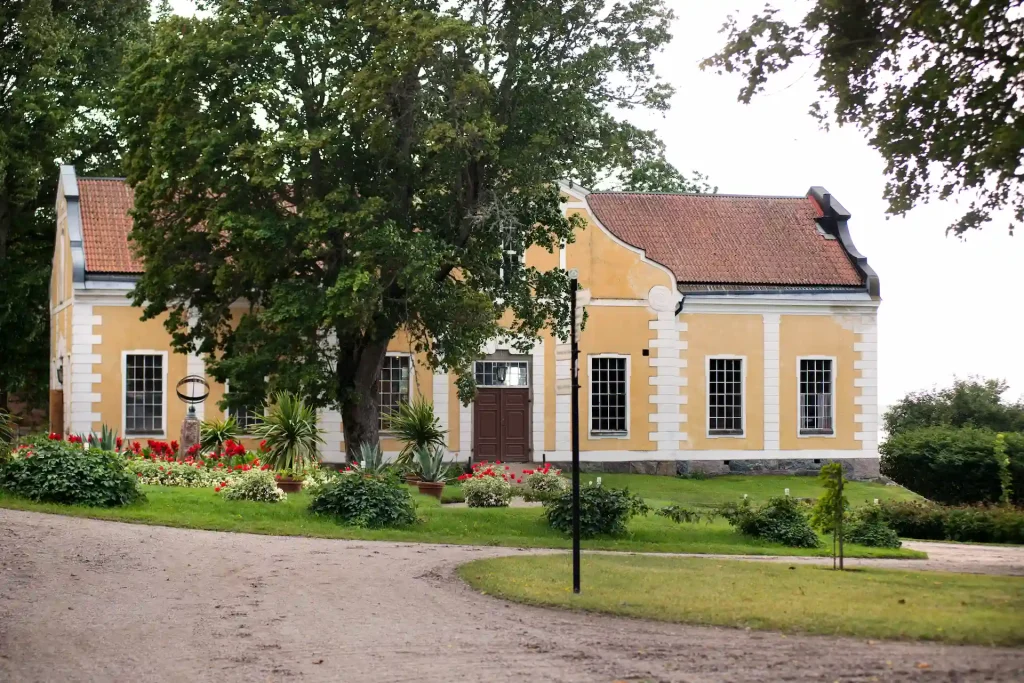 Gul byggnad med vita knutar och svart tak, omgiven av grönskande trädgård och ett träd i framgrunden.