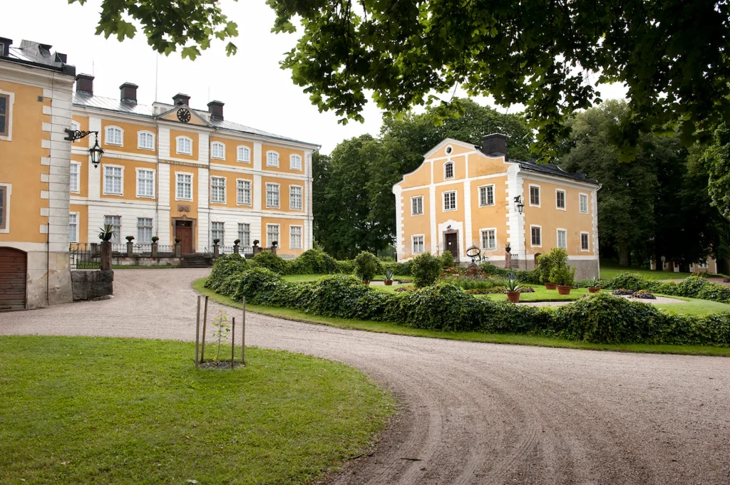 Tre gula herrgårdsbyggnader med en slingrande grusgång och grön plantering.