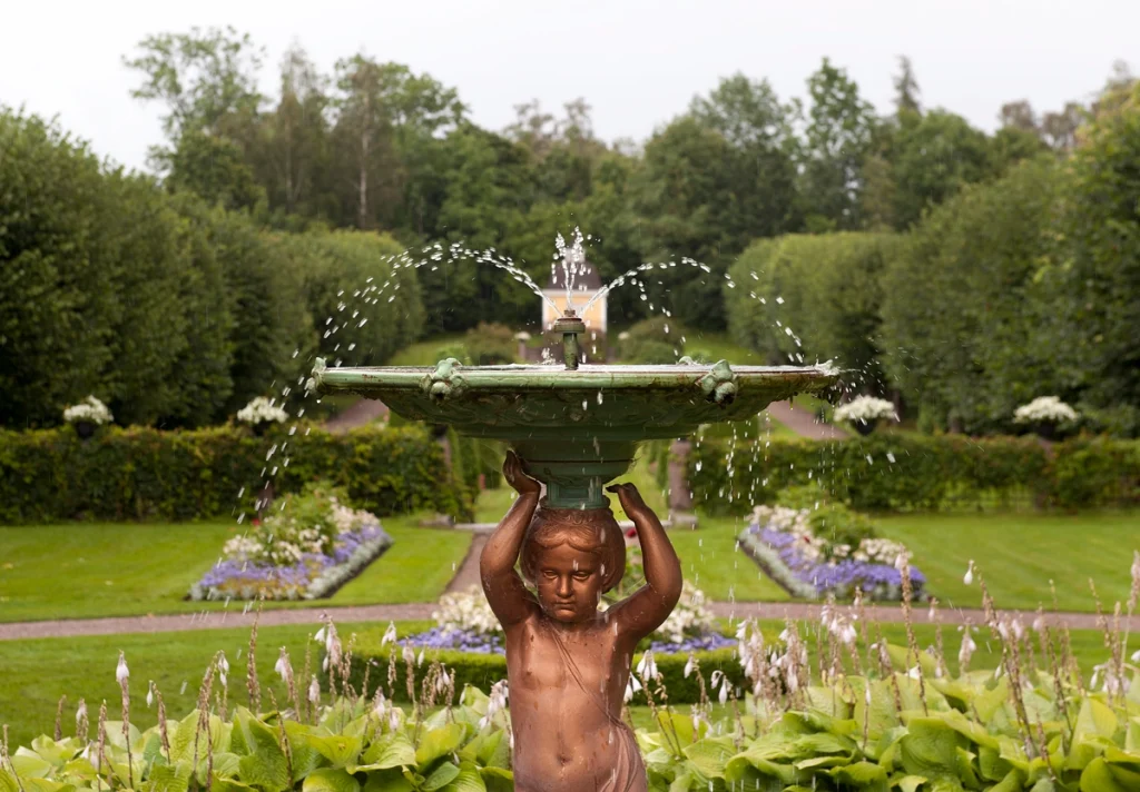 En fontän i förgrunden med en herrgårdspark med rabatter, häckar och ett lusthus i bakgrunden.