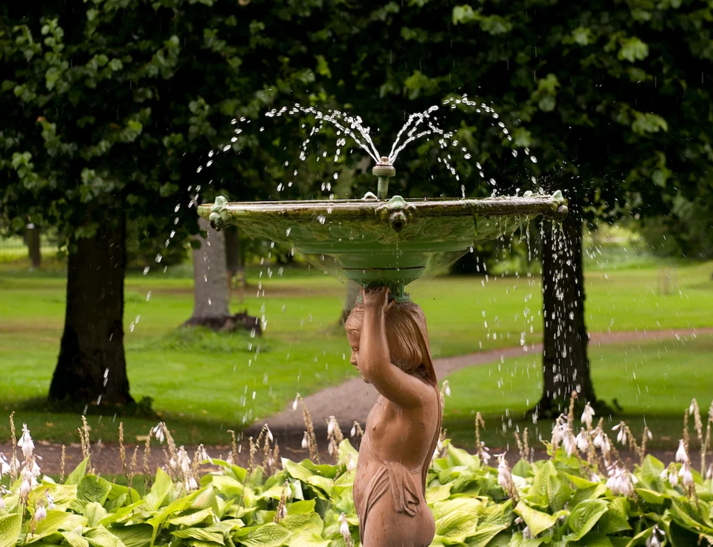 En park med en fontän i form av en amorin, runtom funkior och stora lindar i bakgrunden.