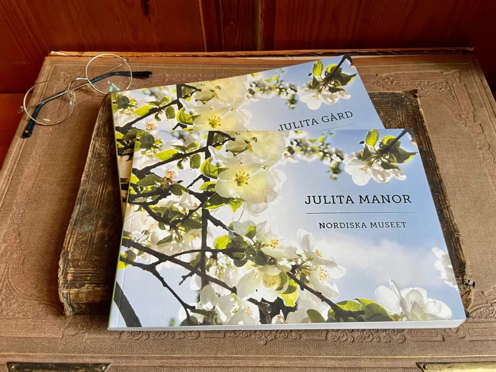 Två kataloger med blommande äppelträd ligger på ett äldre skrivbord med glasögon bredvid sig, träpanel i bakgrunden.