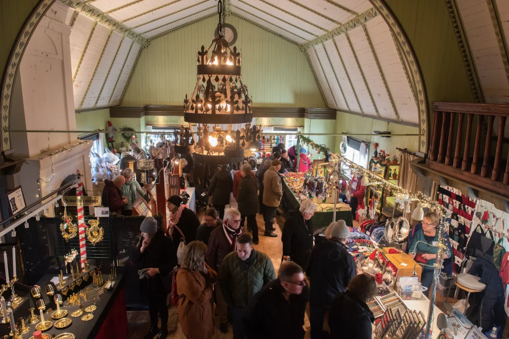 Julmarknadsstånd inne i Skansenmuseet på Julita gård, många människor i ytterkläder som tittar på pynt och dekorationer.