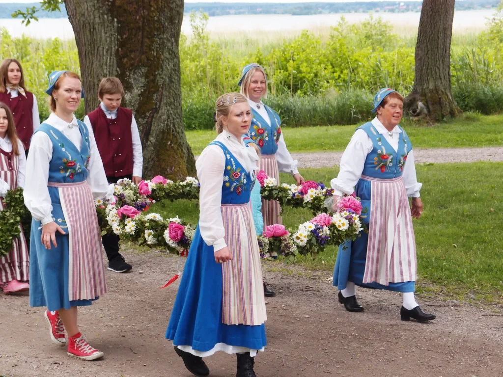 Sju folkdräktklädda personer i olika åldrar bär fram en klädd midsommarstång på en grusväg, en sjö i bakgrunden.