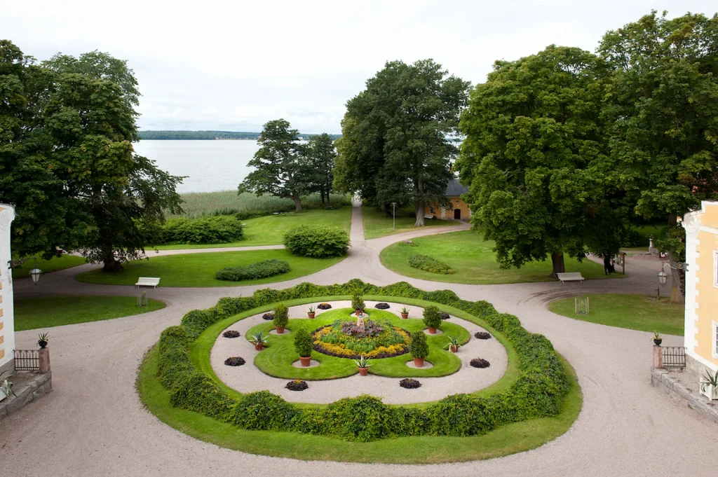 En rund plantering med krukor och slingrande vildvin, runtom grusgångar, parkträd och i fonden en sjö.