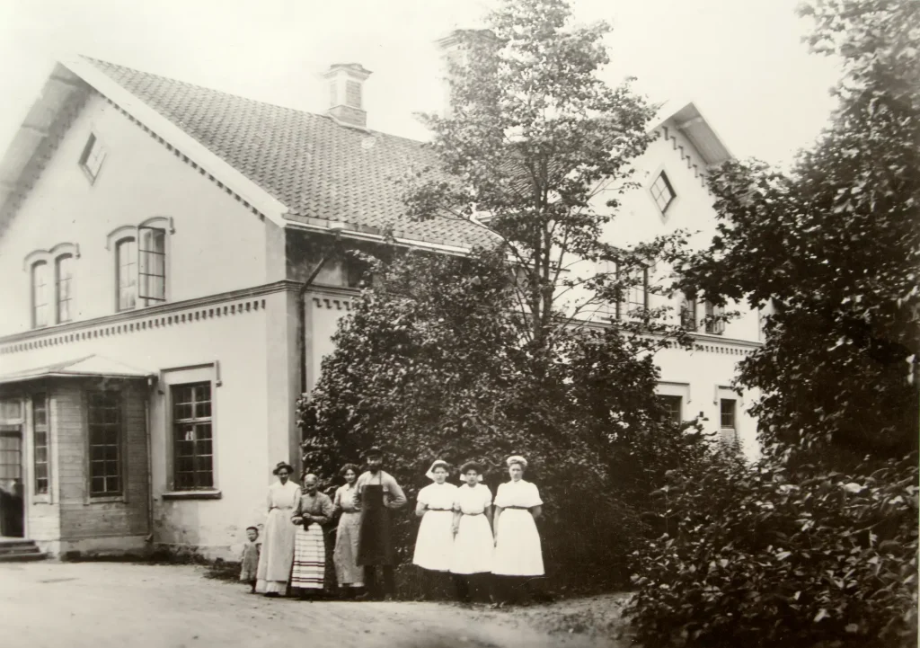Ett äldre svartvitt fotografi på ett äldre hus med stuckatur i knutarna, omgivet av träd och buskar på höger sida. Framför huset står tre flickor i vita klänningar och till vänster om flickorna står fyra personer i olika förkläden.