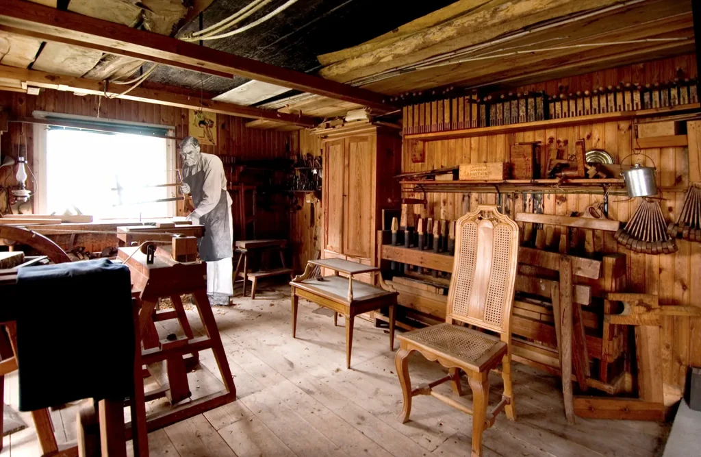 En verkstad i träutstyrsel med verktyg på väggarna, en arbetsbänk till vänster med en pappfigur på en man med en såg bakom. Mer centrerat står två möbler, en stol och ett lågt bord.