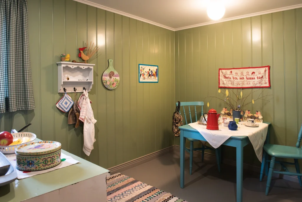 Ett kök i en lekstugan med gröna pärlspåntväggar, blått bord och stolar och trasmatta på golvet.
