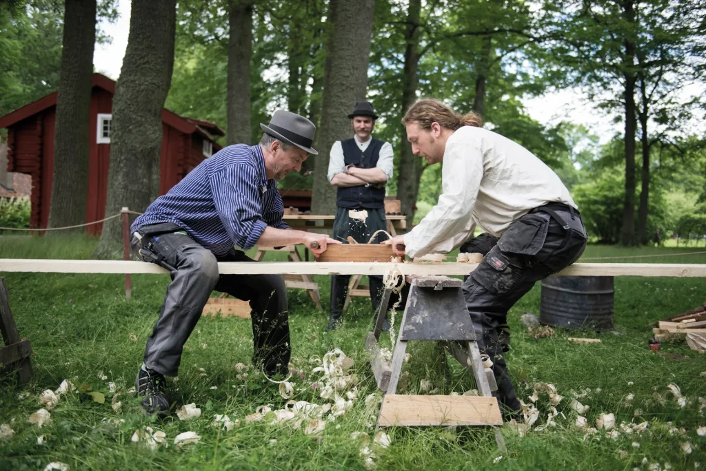 Två män står mitt emot varandra utomhus och täljer medan en tredje man står lite längre bort och tittar på. I bakgrunden syns natur, himmel och en röd stuga.