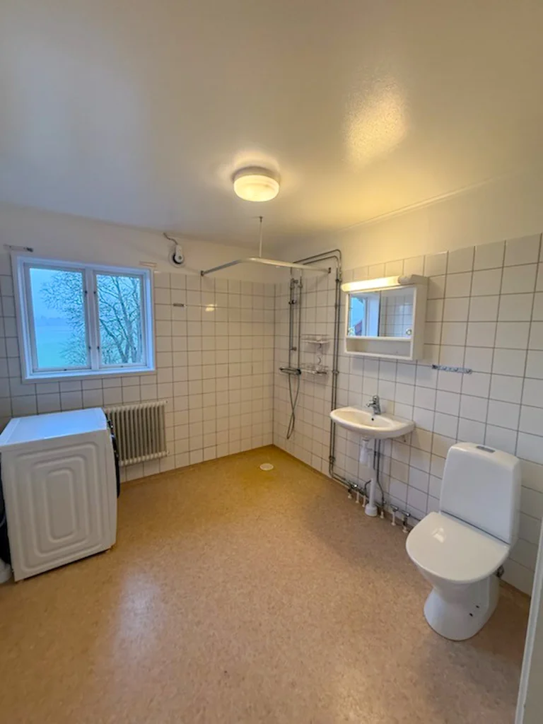 Ett badrum med plastmatta, vitt kakel på väggarna, wc, dusch, tvättmaskin och stort fönster.