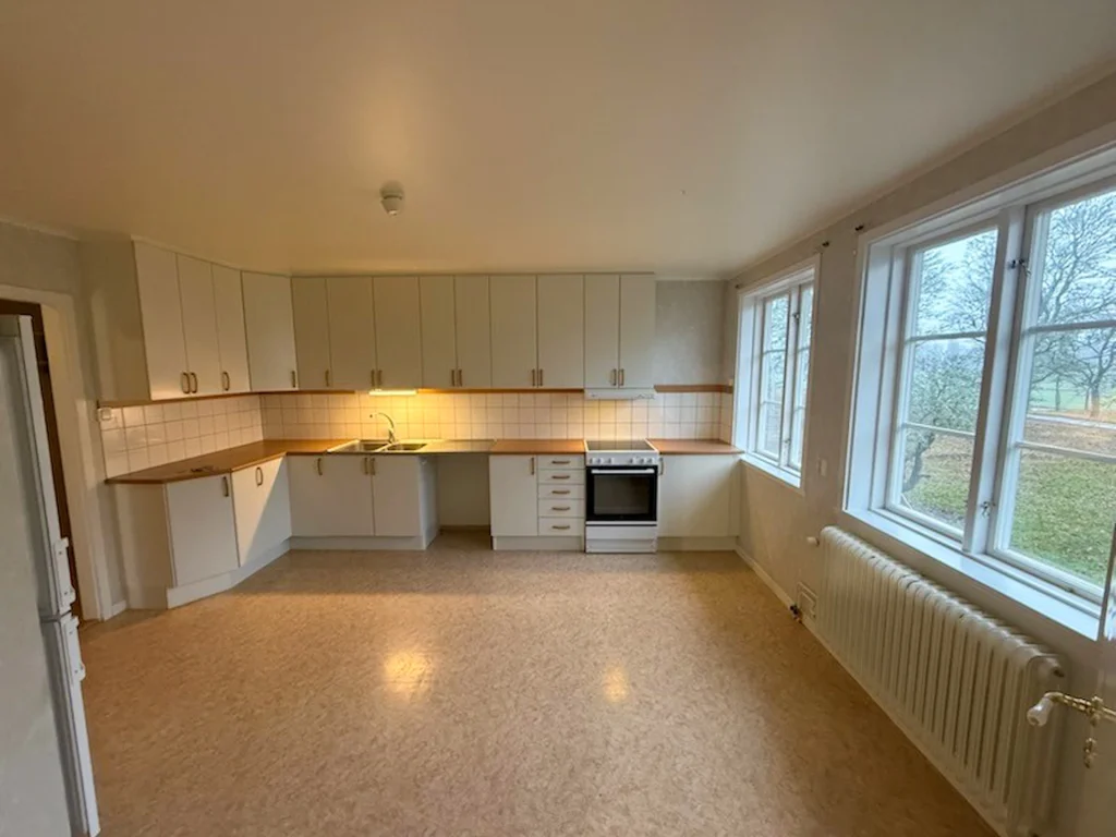 Ett omöblerat stort rymlig kök med vita luckor, bönksiva i trä och kork-och-plastmatta.