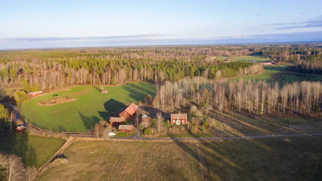 Flygfoto över ett rött torp med lada som ligger vid en grusväg, åkrar, skog och fält omkring.