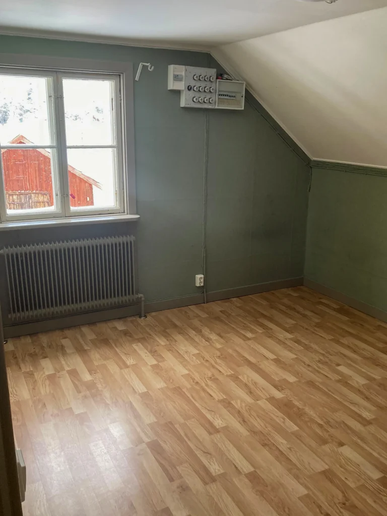 Ett tomt sovrum med parkettgolv, snedtak och fönster, grågrönmålade väggar..