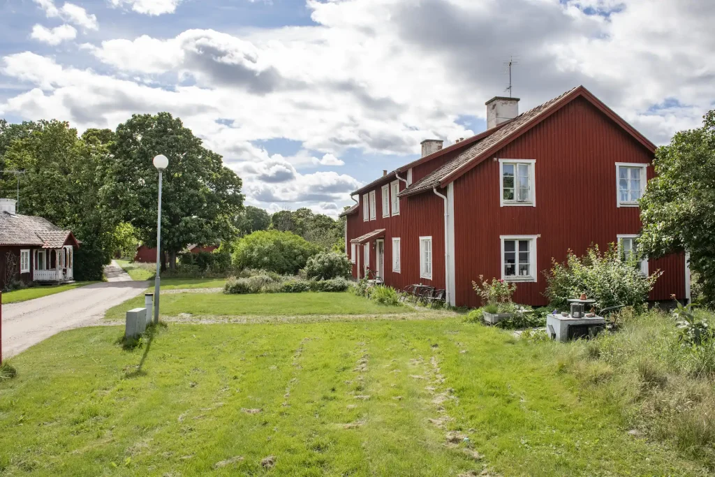 En faluröd länga med vita knutar sedd från kortsidan, gräsmatta buskar och utemöbler framför huset