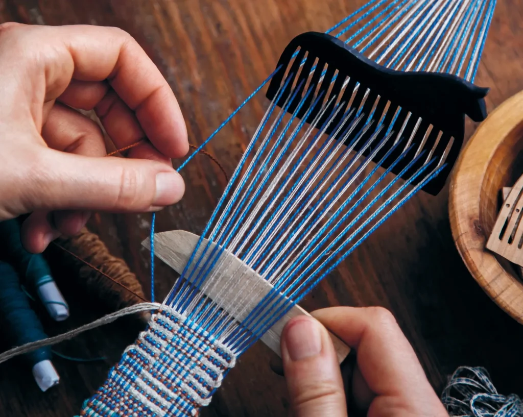 Närbild av händer som väver blå tråd i en liten bandgrind