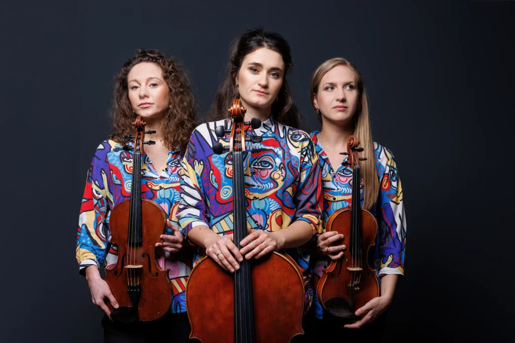 Tre unga kvinnor i mönstrade skjortor håller i varsitt stråkinstrument